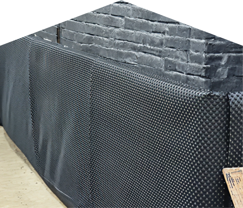 Exterior basement waterproofing completed by Furnace Guys, showing a sealed foundation wall with dimpled waterproof membrane partially installed on a brick surface, preventing moisture infiltration.