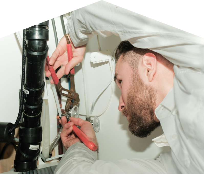 Furnace Guys plumbing specialist uses hand tools to tighten pipe fittings beneath a sink during a repair, with visible wiring and drainage lines in the background. Furnace Guys plumbing specialist uses hand tools to tighten pipe fittings beneath a sink during a repair, with visible wiring and drainage lines in the background.