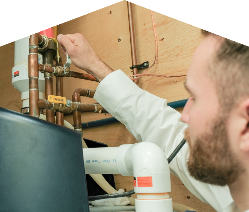 Furnace Guys water heater specialist tightens a valve on copper piping during a water heater installation, with exposed wood paneling and wiring in the background. Furnace Guys water heater specialist tightens a valve on copper piping during a water heater installation, with exposed wood paneling and wiring in the background.